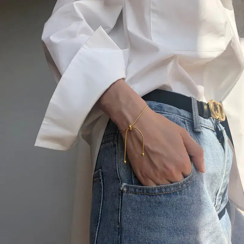 Person wearing a white shirt and blue jeans with a gold bracelet on a plain background
